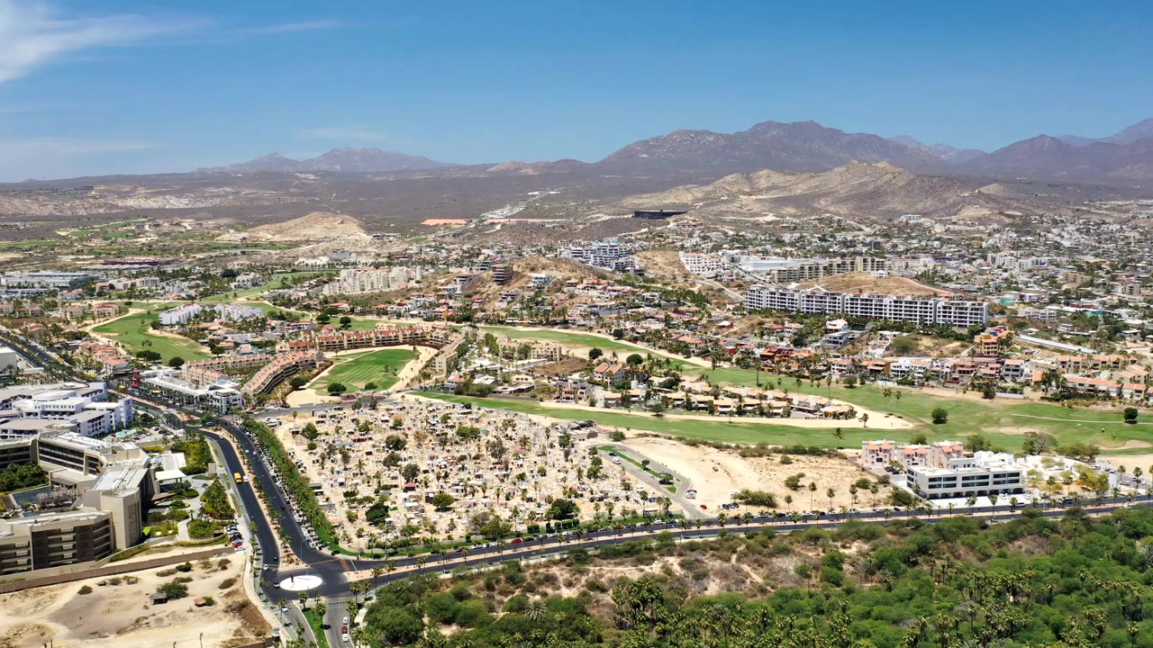 Wide aerial view over Los Cabos, Mexico, with foreground roads and tiny cars leading toward golf greens, low buildings, desert lots, and distant mountains, with no recognizable people visible.