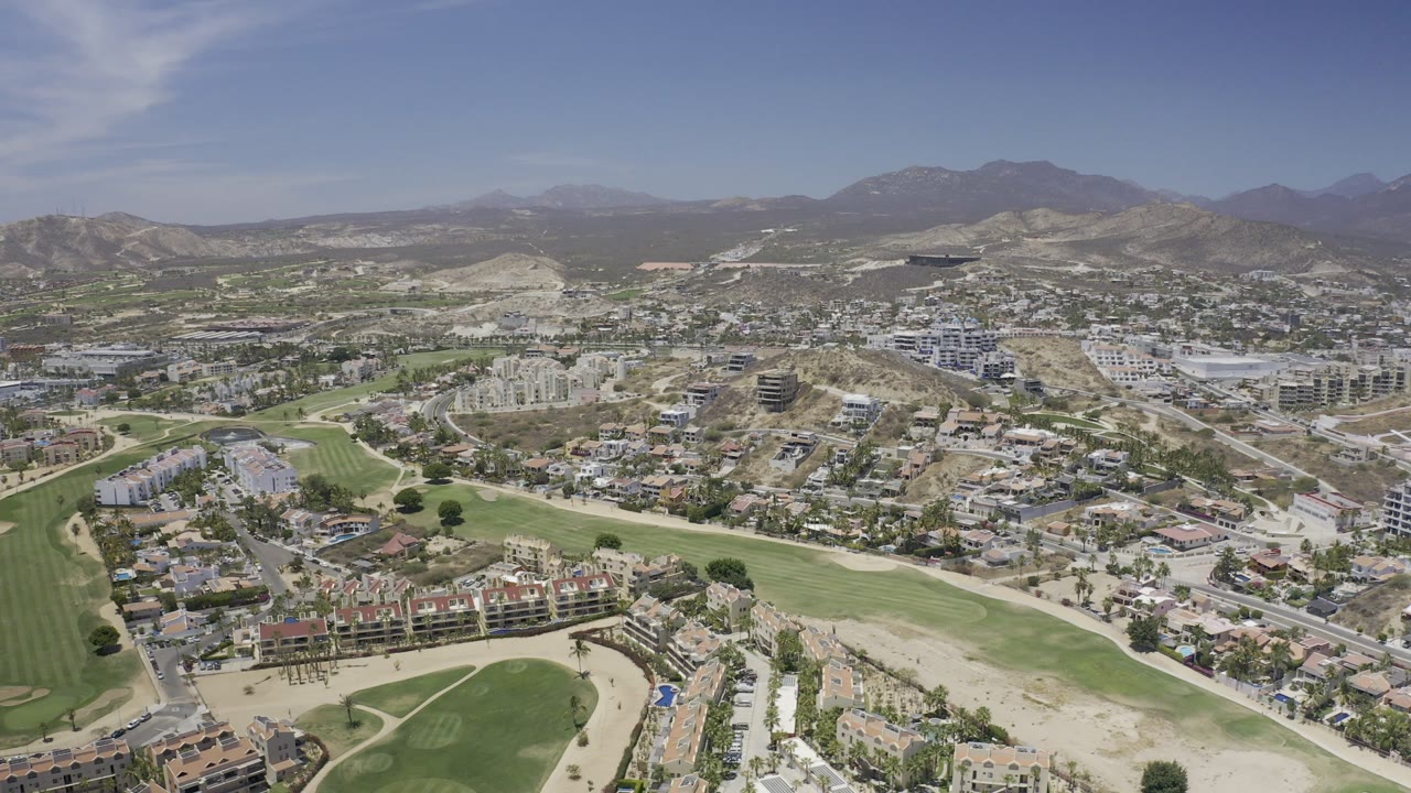 High aerial view over Los Cabos, Mexico, with suburban homes, golf-course greens, roads, dry hills, distant mountains, blue sky, and no visible people.