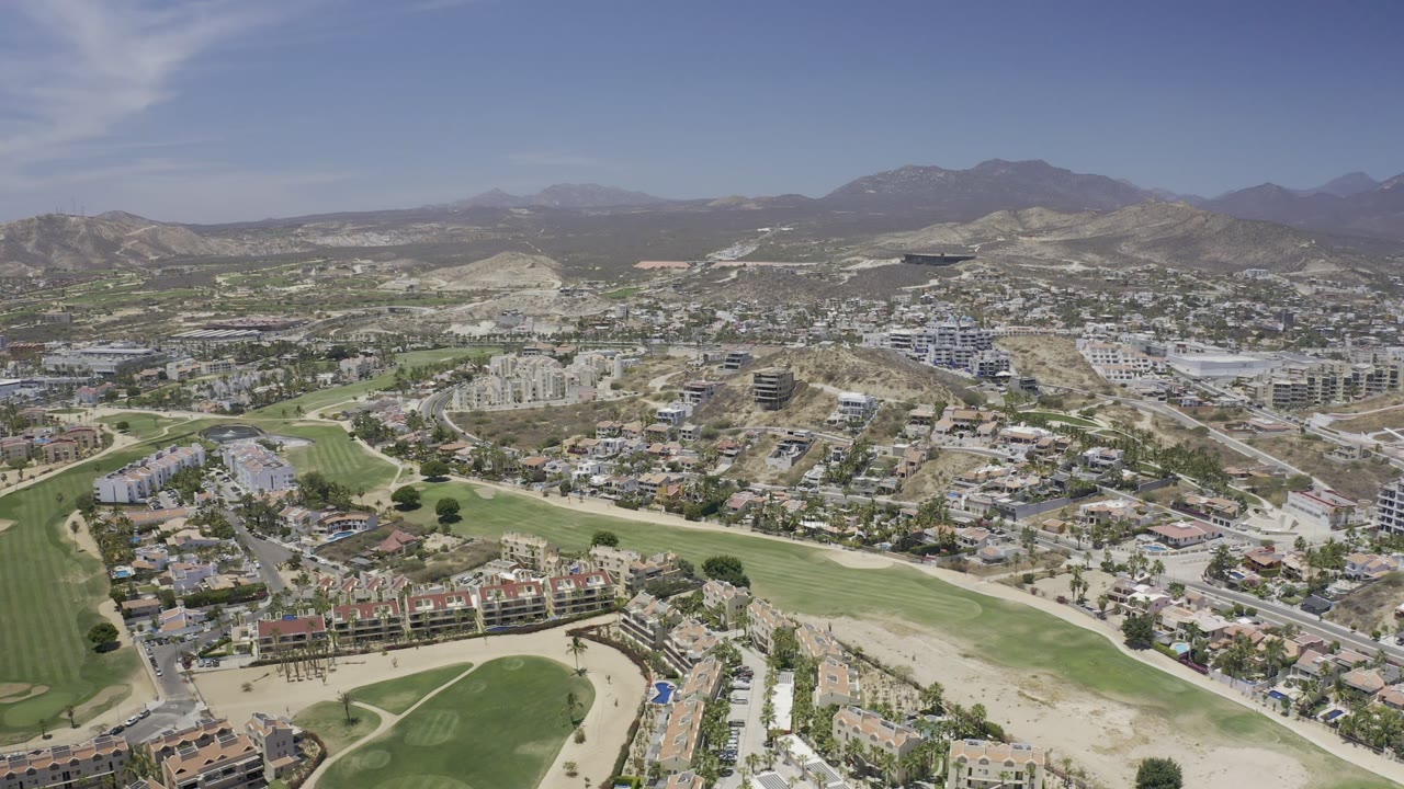 Aerial frame in Los Cabos, Mexico, looking across fairways, clusters of homes, wider roads, construction areas, arid hills, distant mountains, and no visible people.