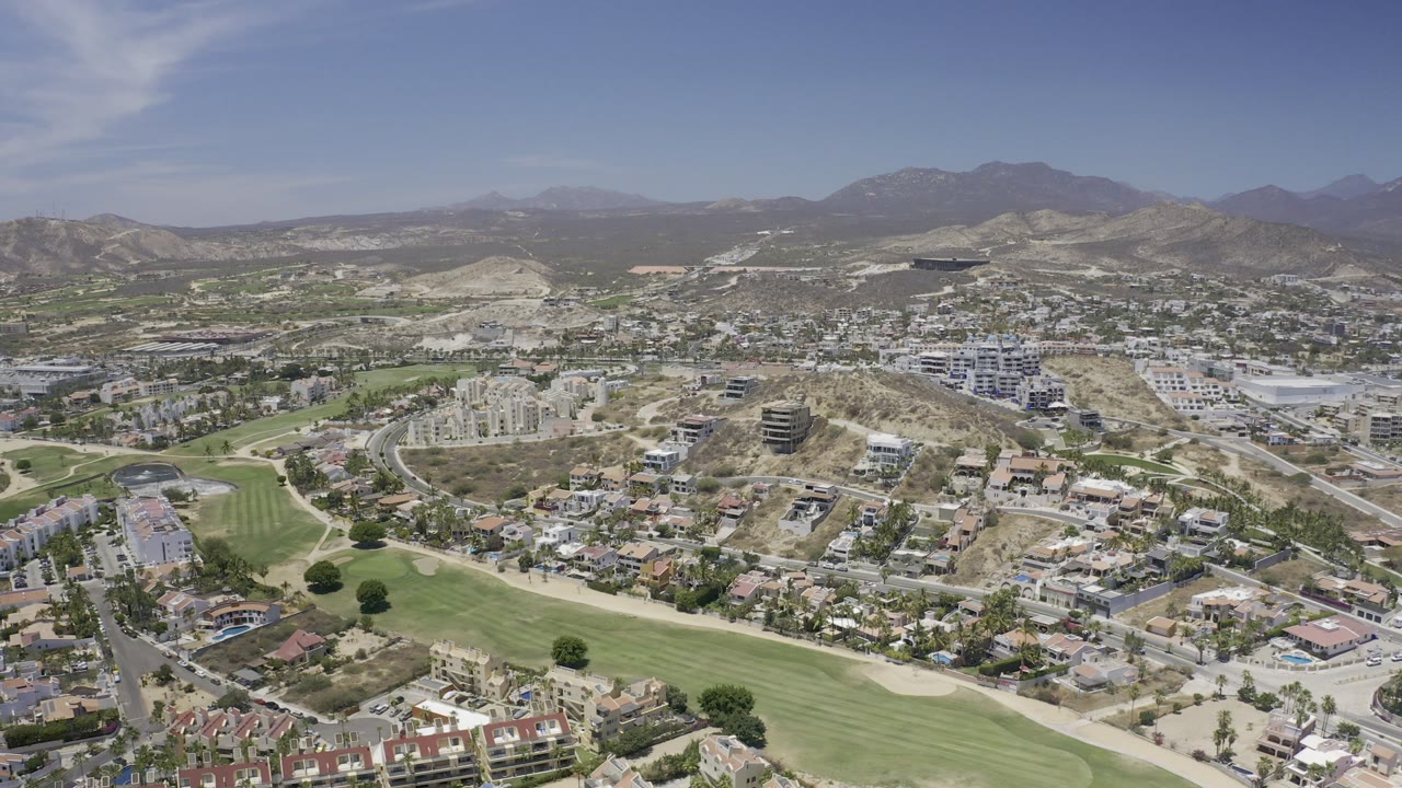 Wide aerial frame over Los Cabos with golf fairways in the foreground, residential streets beyond, dry hills and mountains on the horizon, and no visible people.