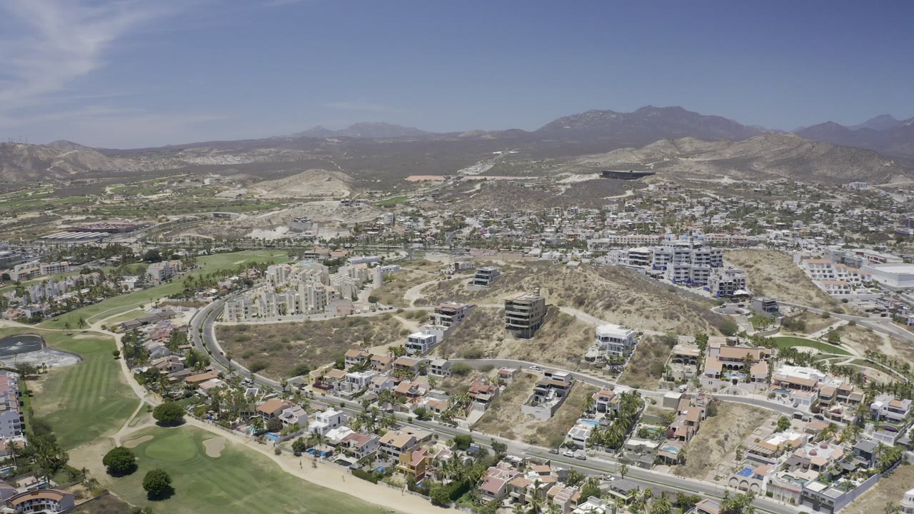 Opening aerial frame over Los Cabos with golf greens at the lower left, hillside homes and condos across the center, mountains on the horizon, and no visible people.