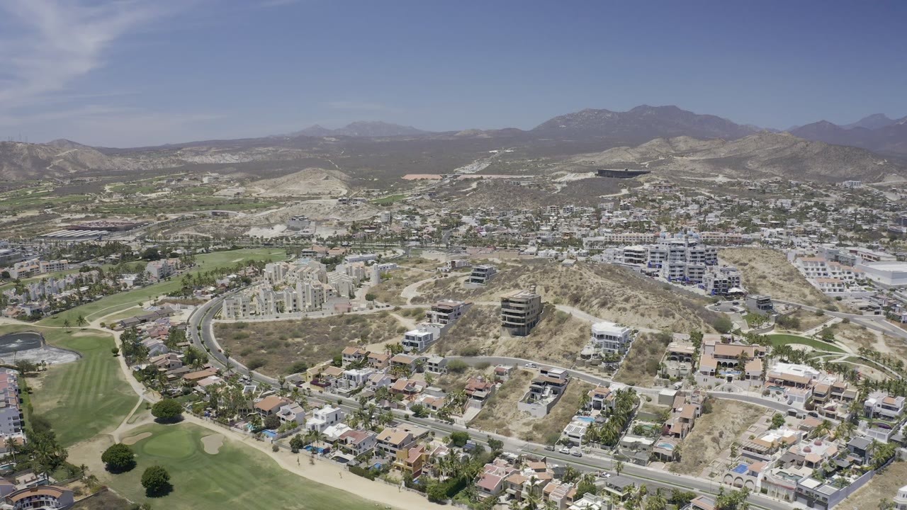 Aerial frame of Los Cabos showing a curved road through residential buildings, green fairways at left, dry hills in the middle distance, and no visible people.