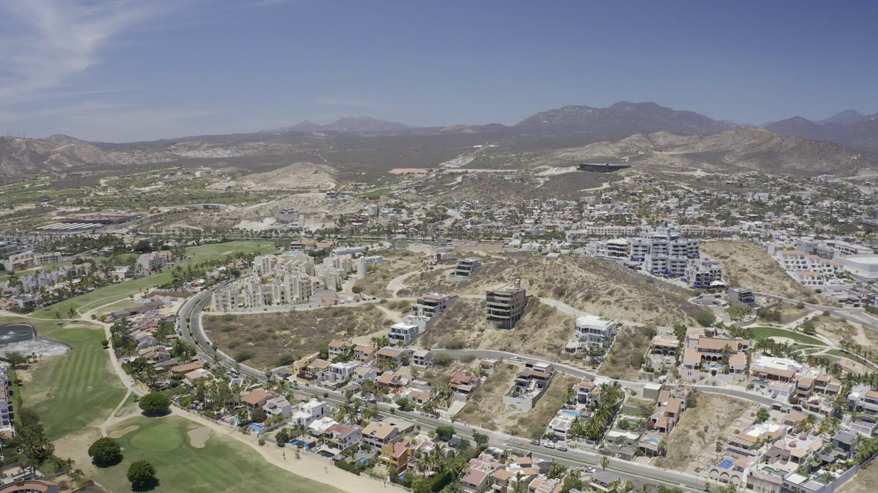 Wide aerial of Los Cabos buildings, fairways, dry hills, and mountains.