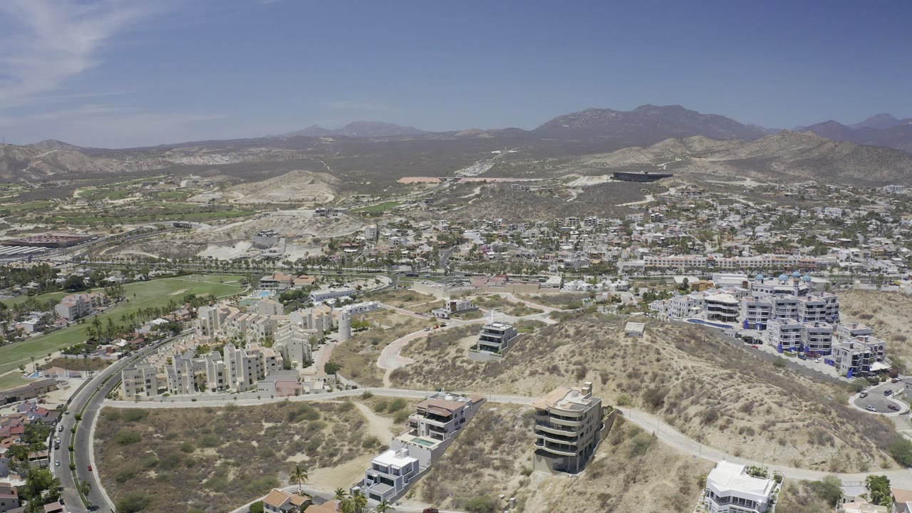 Aerial view over dry hills, roads, buildings, greenery, and distant mountains.