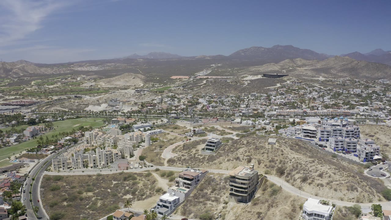 Aerial view in Los Cabos, Mexico, looking across tan hills, light-colored buildings, winding roads, green grounds, a spread-out town area, and no visible people.