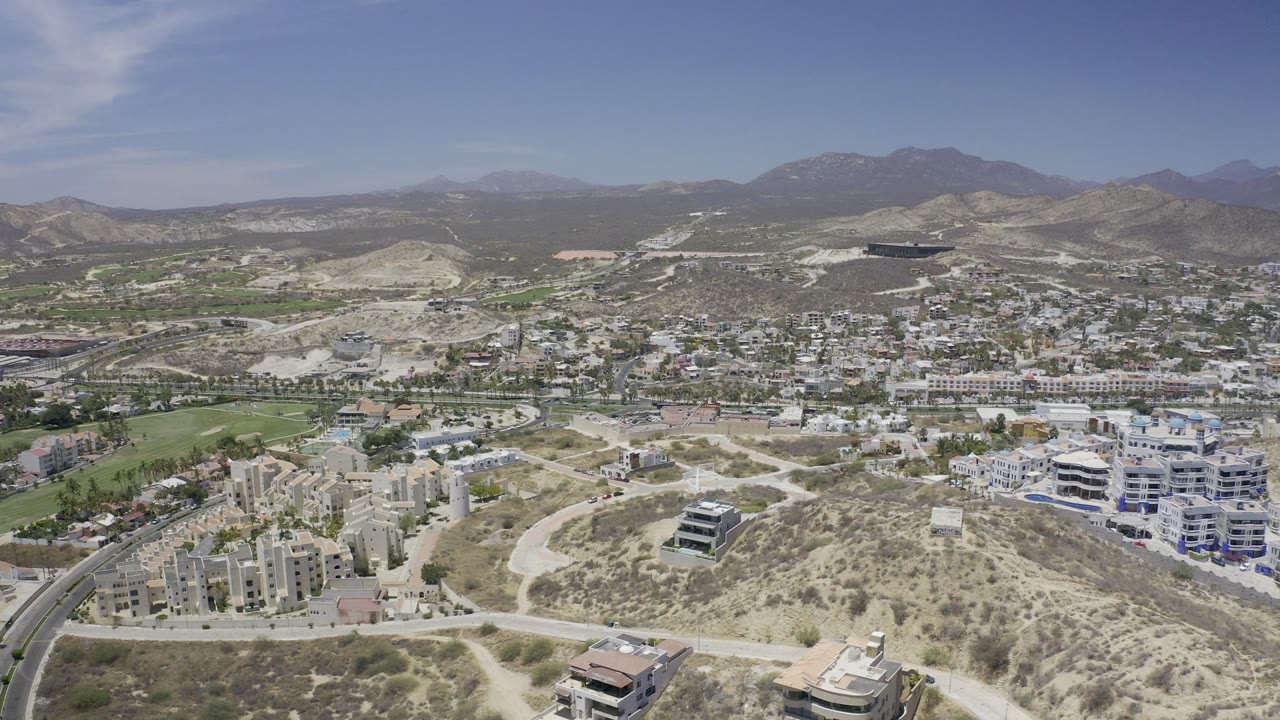 Wide aerial view over Los Cabos, Mexico, showing scrub hills, winding roads, apartment buildings, green grounds, distant mountains, and no visible people.