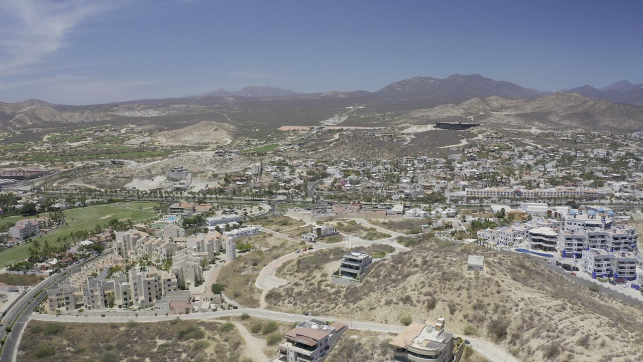 Aerial frame over Los Cabos with a pale dirt road crossing the foreground slope, dense white buildings in the midground, mountain ridges behind, and no visible people.