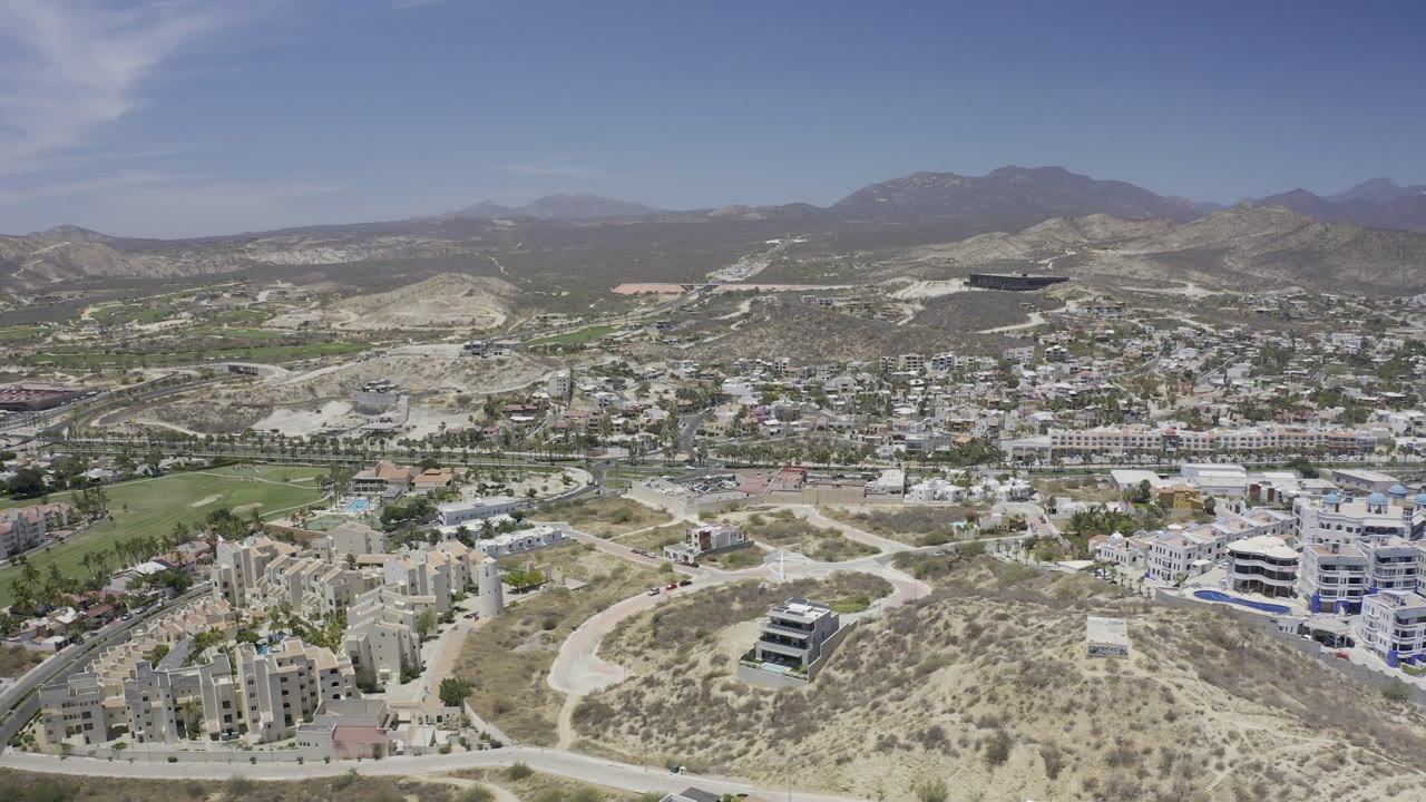 High aerial view in Los Cabos with pale hills in the foreground, white buildings and roads across the middle, green landscaped areas, mountains, and no visible people.