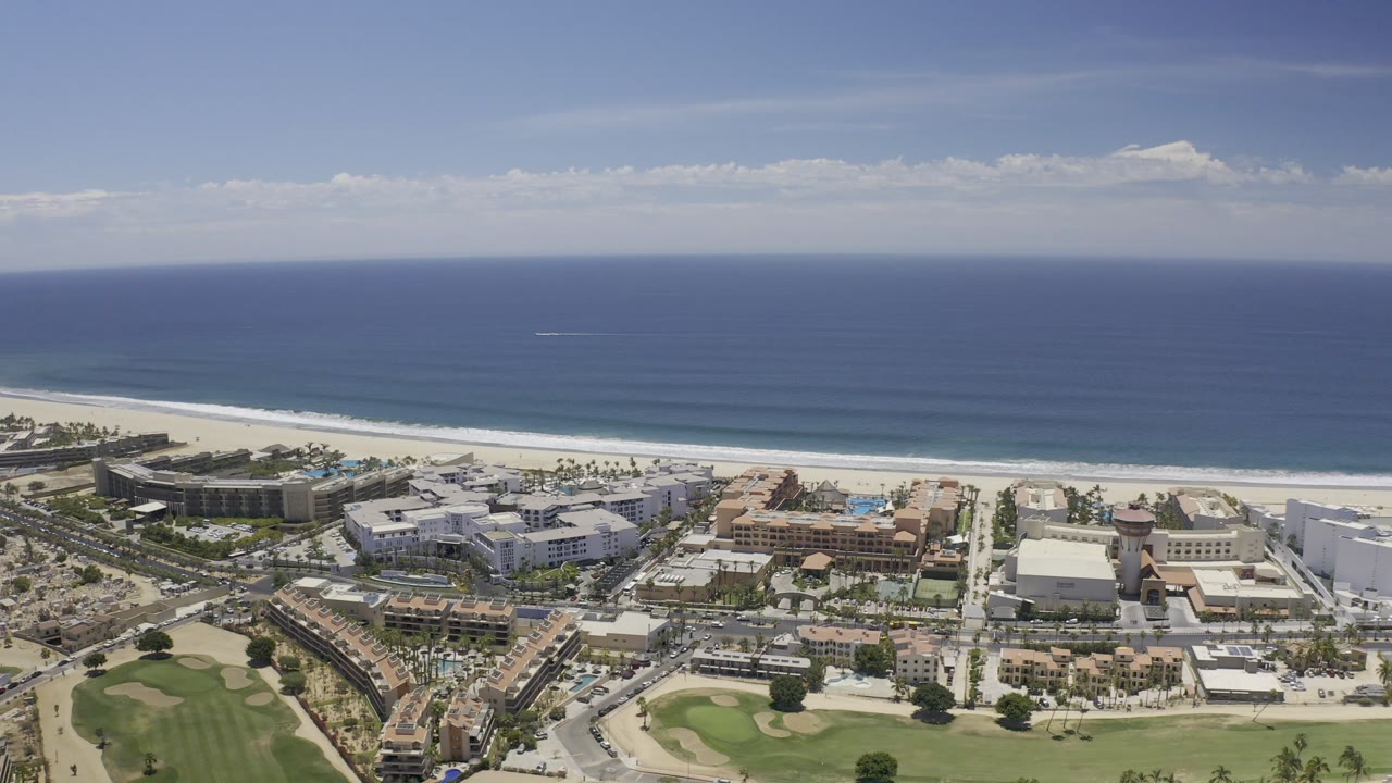 High aerial view over Los Cabos, Mexico, with blue ocean, pale beach, beachfront buildings, roads, and green golf course foreground, no visible people.