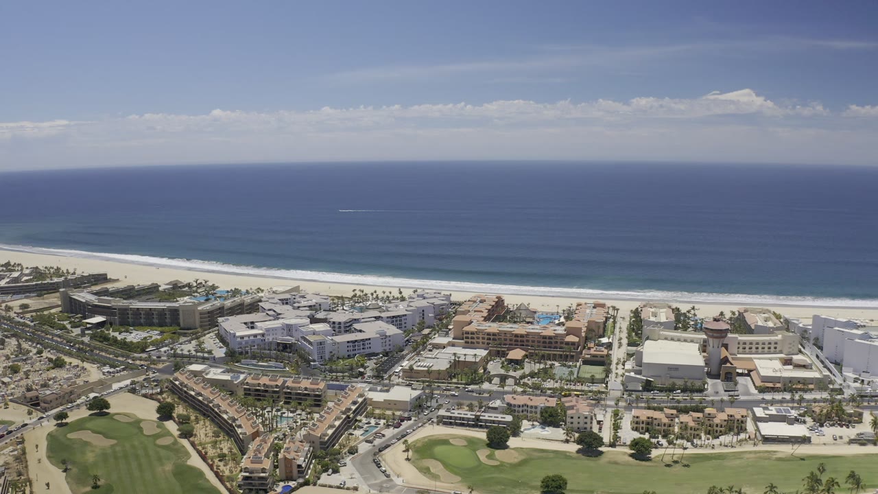 Aerial Los Cabos coastline view with long surf bands, beachfront development, a straight road to the beach, and golf greens in the lower frame, no visible people.