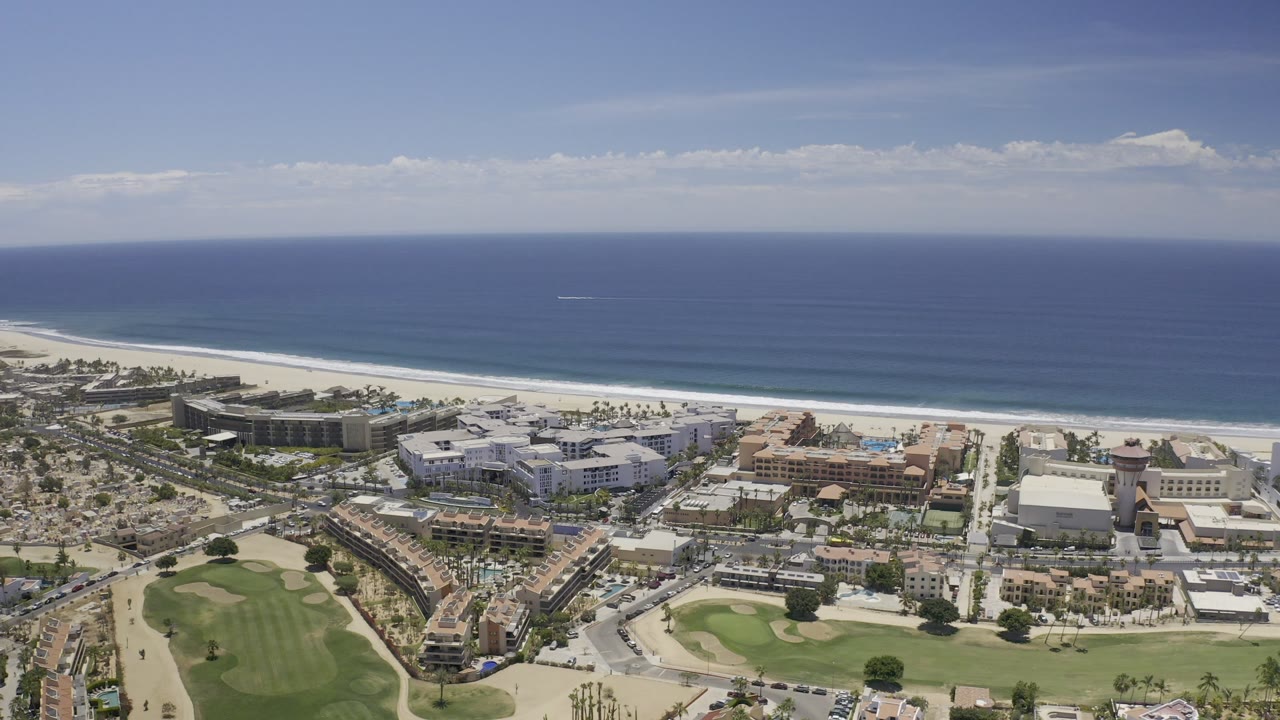 Opening aerial frame over Los Cabos, Mexico with beachfront buildings, green lawns, a wide sand beach, blue ocean, and no visible people.