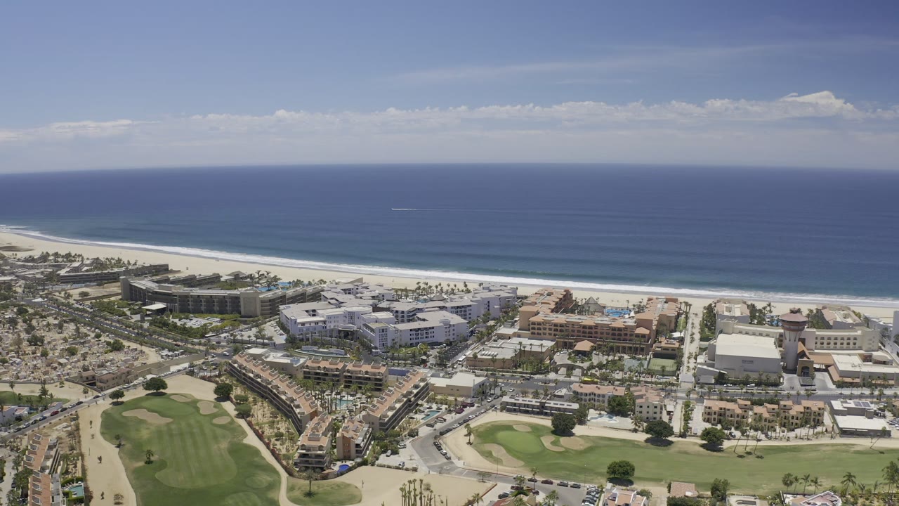 Second aerial frame in Los Cabos, Mexico showing the shoreline stretched across the image, with hotels or large buildings, homes, roads, and no visible people.
