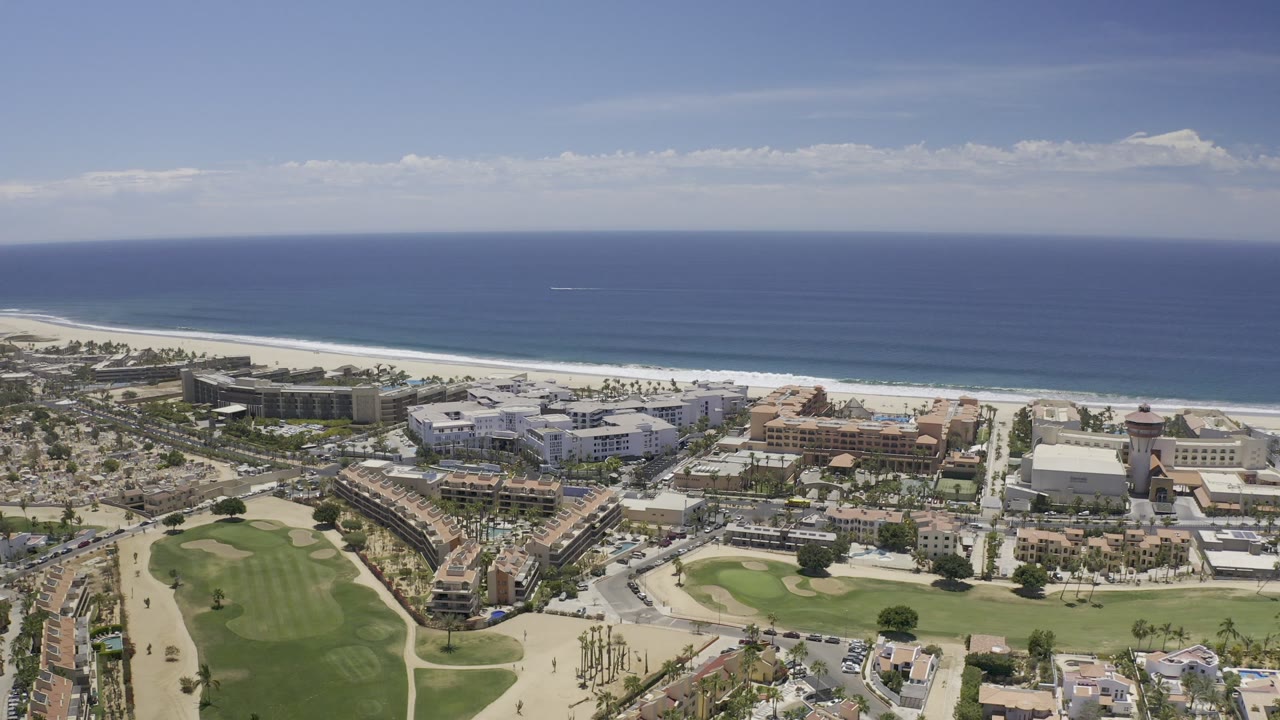 Opening aerial frame over Los Cabos, Mexico, showing blue ocean, a white surf line, beachfront buildings, paved roads, green lawns, sandy paths, and no visible people.