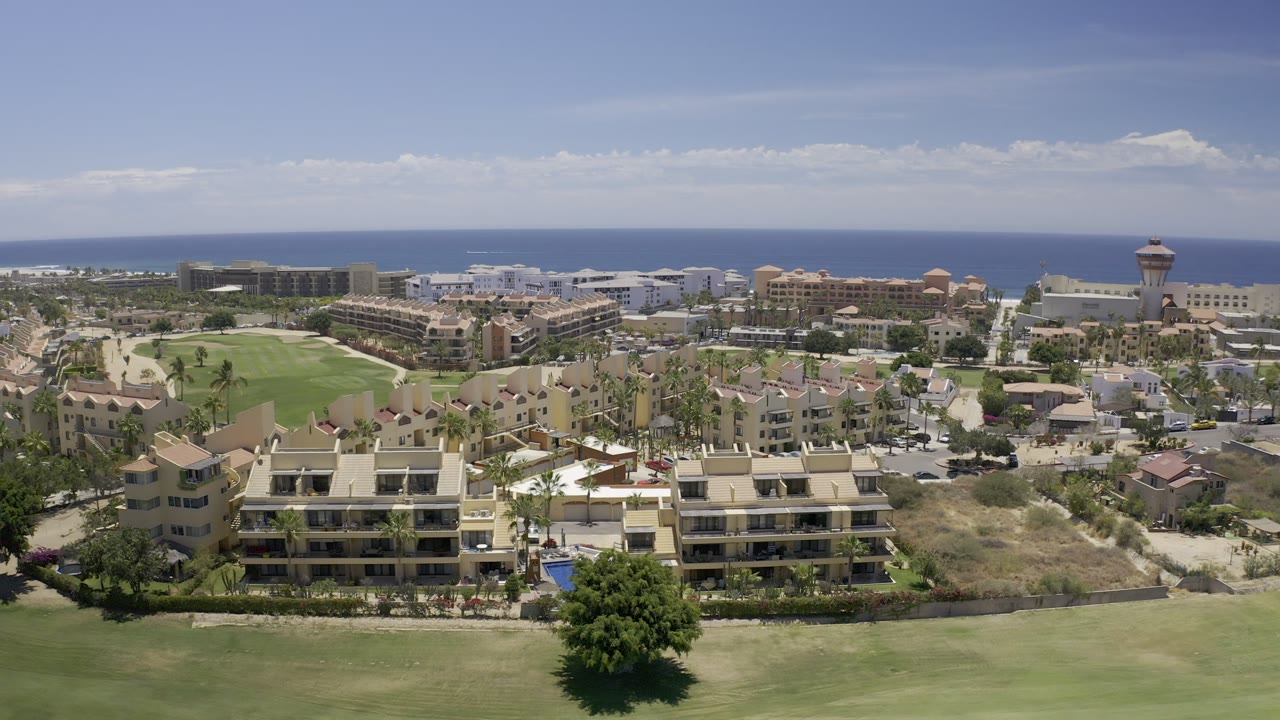 Los Cabos aerial frame showing more of the fairway curve at the bottom, rows of light-colored buildings, a tower at right, blue ocean, and no visible people.