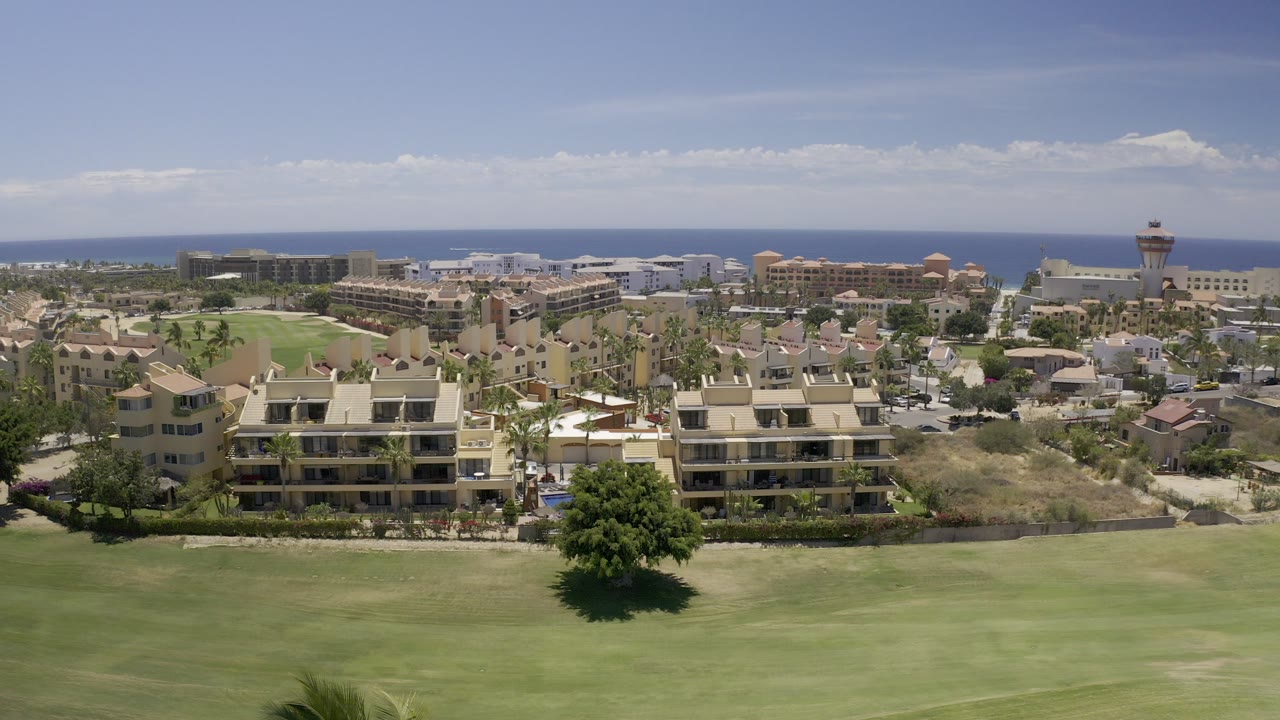Opening frame of Los Cabos, Mexico from an elevated view, with a wide green lawn in front, tan residential buildings in the middle distance, coastal development near the water, blue sky above, and no visible people.