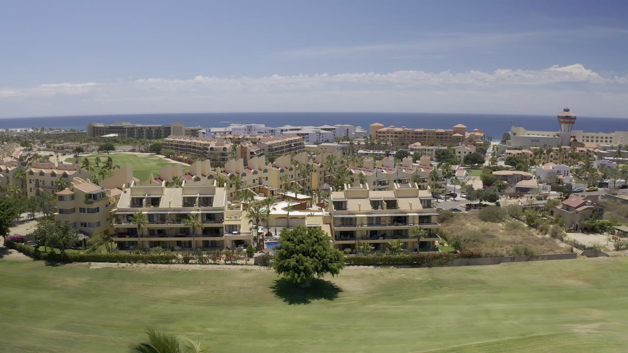 Los Cabos aerial frame with the lawn filling the lower half, palms and beige residences across the midground, larger coastal buildings near the horizon, a tower-like structure to the right, and no visible people.