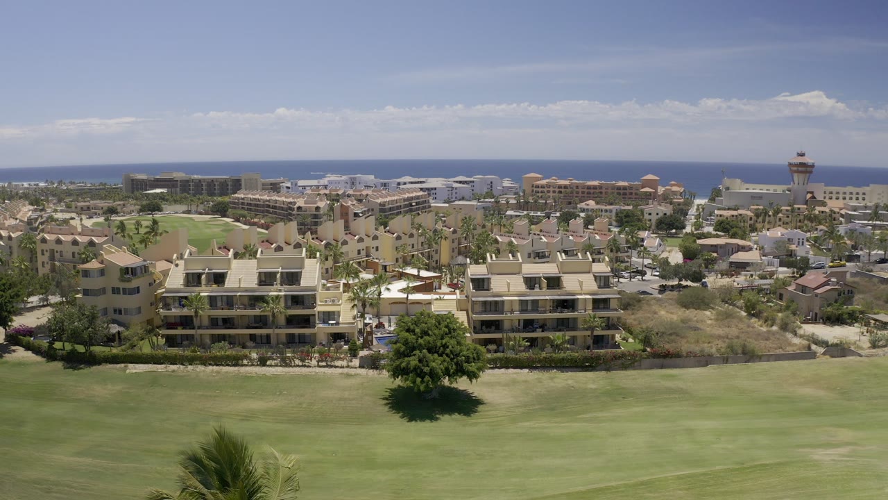 Wide elevated view of palm-lined residences with a green foreground lawn and blue ocean in the distance.