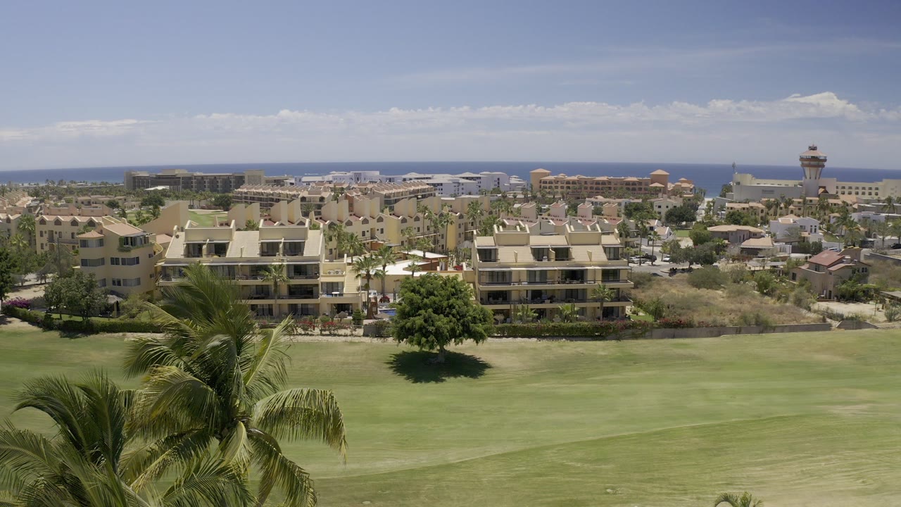 Opening Los Cabos frame with a wide elevated view over a green golf-course fairway, low-rise buildings, distant sea horizon, pale clouds, and no visible people.