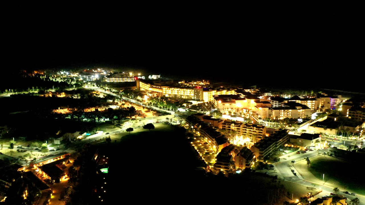 Los Cabos night aerial with a bright diagonal road through the center, warm building lights on the right, darker open ground on the left, and no visible people.