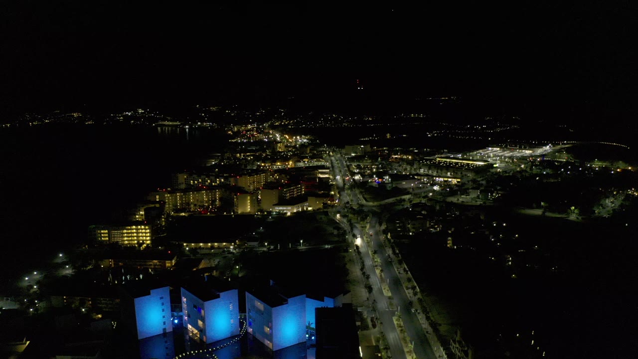 Opening frame of Los Cabos at night from above, with blue-lit buildings in the foreground, bright roads through the center, distant shoreline lights, and no visible people.