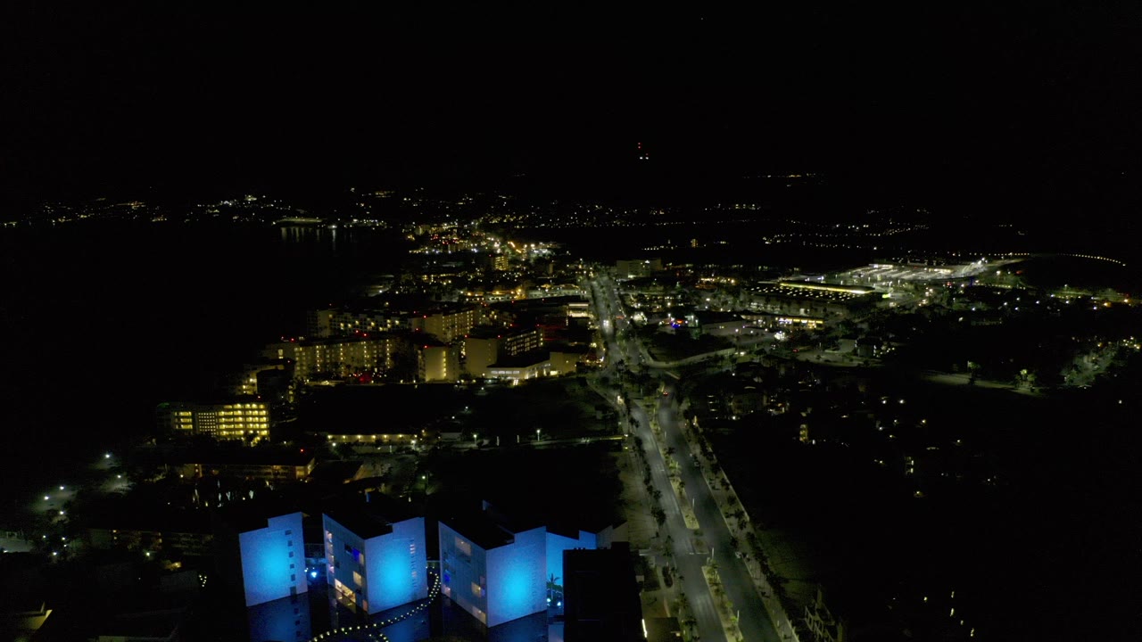 Opening night aerial frame over Los Cabos, Mexico, with blue-lit foreground buildings, a bright road corridor, scattered distant lights, and no visible people.