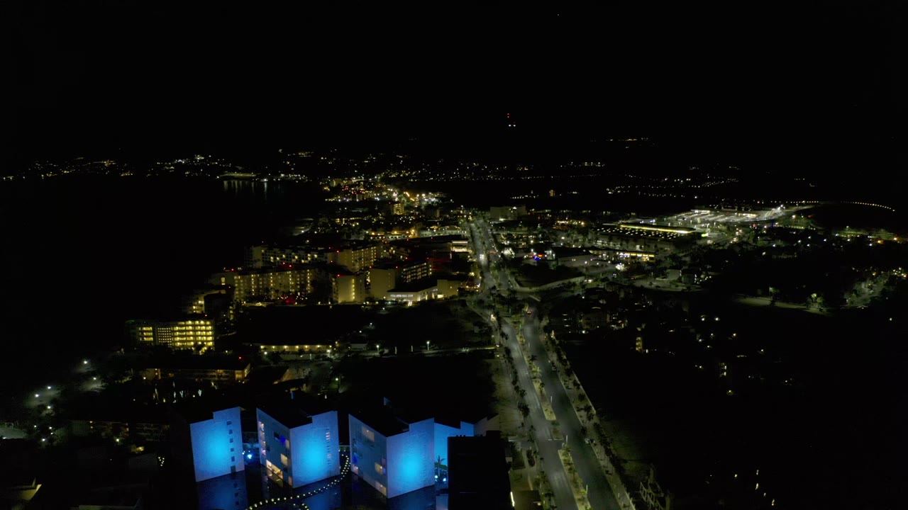Los Cabos night overview with the main illuminated street running through the center, blue foreground structures below, distant buildings, and no visible people.