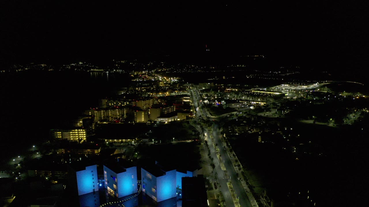 Night aerial view of city lights, roads, and blue-lit foreground buildings.