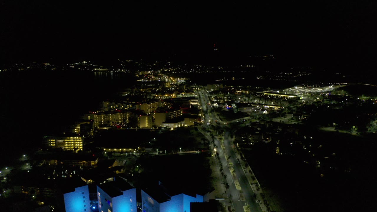 Opening frame of Los Cabos, Mexico, at night with a bright highway crossing the center, lit buildings on the left, distant lights on the horizon, and no visible people.
