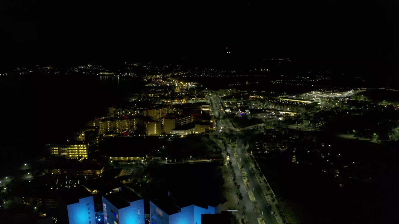Los Cabos, Mexico, night aerial with the main road slightly more centered, yellow building lights at left, a bright road junction ahead, and no visible people.