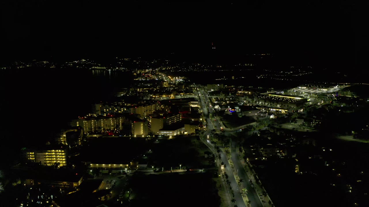 Los Cabos night aerial with yellow-lit building blocks at left, a bright road through the center, scattered horizon lights, and no visible people.