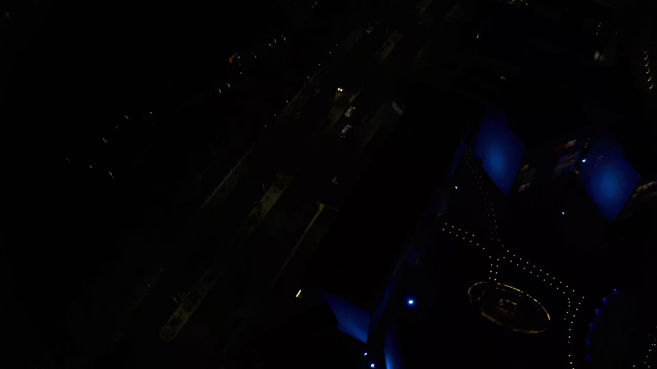 High nighttime aerial frame in Los Cabos, Mexico, showing blue roof lighting, faint roads, small building lights, and no visible people.