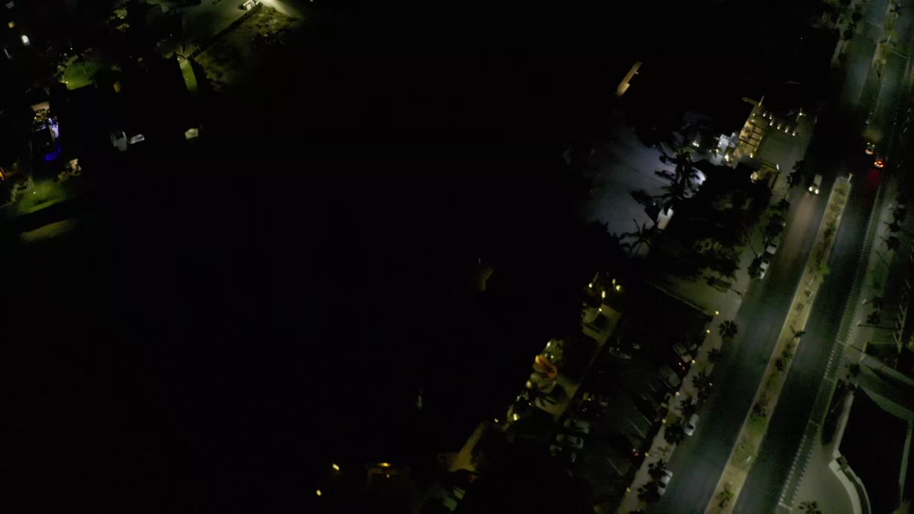 Night aerial frame in Los Cabos showing a diagonal lit roadway, small structures, tree cover, and no visible people.
