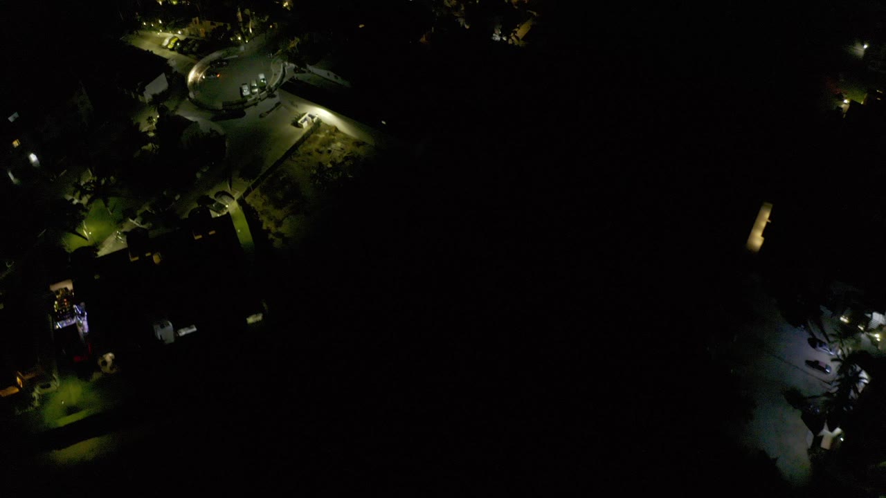 Night aerial view of lit paths and a round structure in a dark town area.