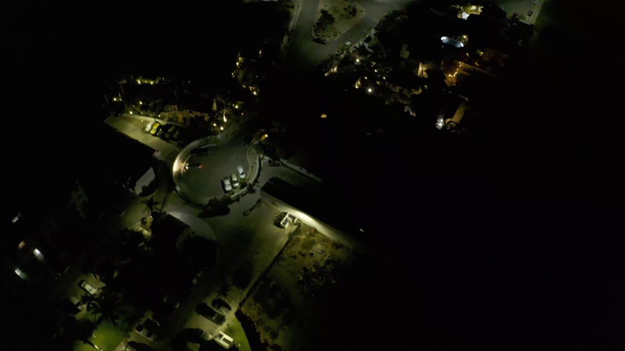 Night aerial view in Los Cabos, Mexico, showing the rounded roadway slightly lower in frame, parked cars near the curve, lit paths, and no visible people.