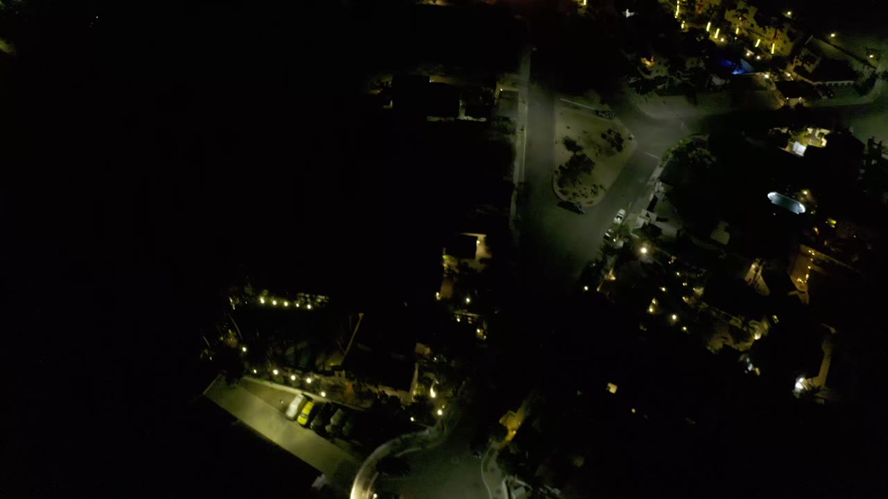 A Los Cabos, Mexico night drone frame shows a bright road junction, scattered homes, trees, parked vehicles, and no visible people.