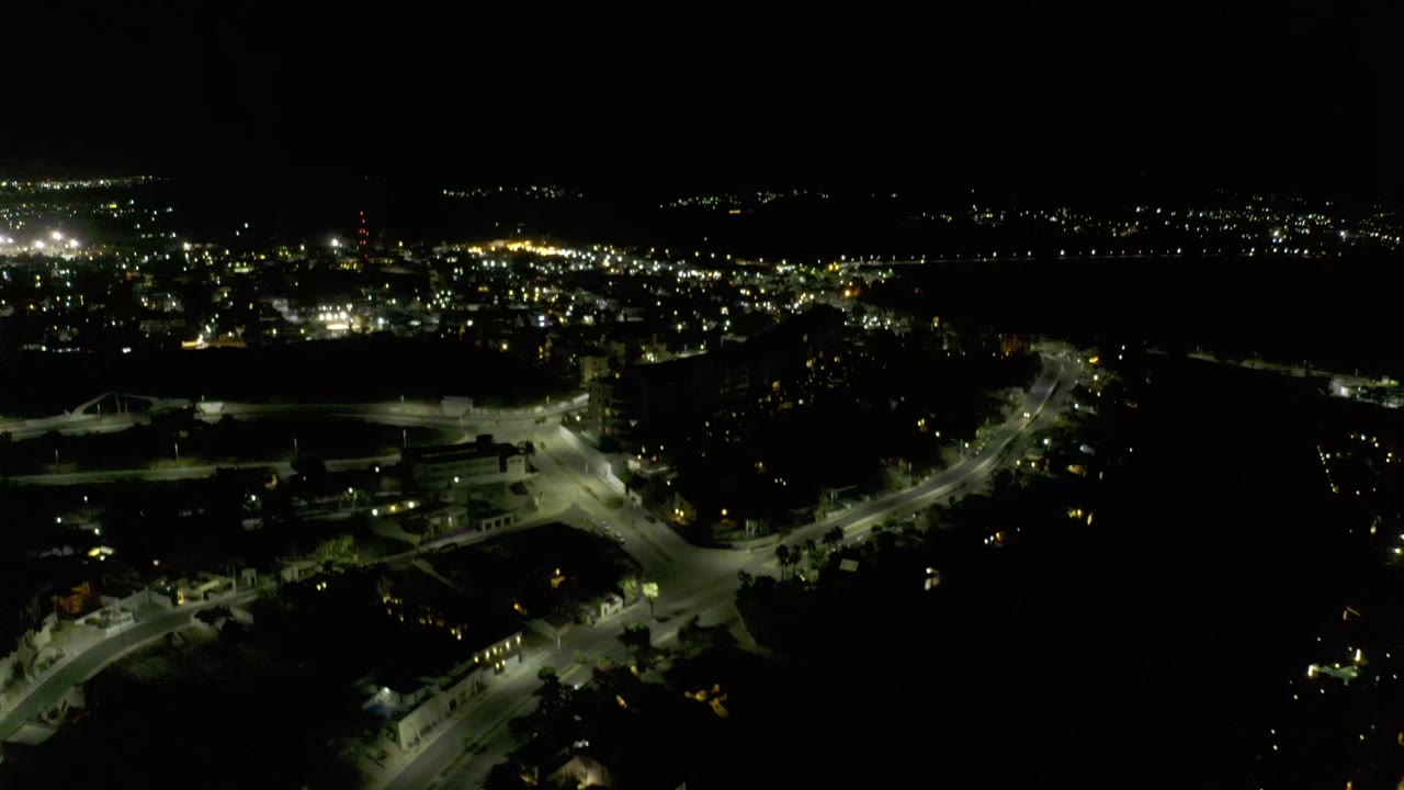 Los Cabos night view with a bright road junction in the lower frame, scattered buildings, distant lights, and no visible people.