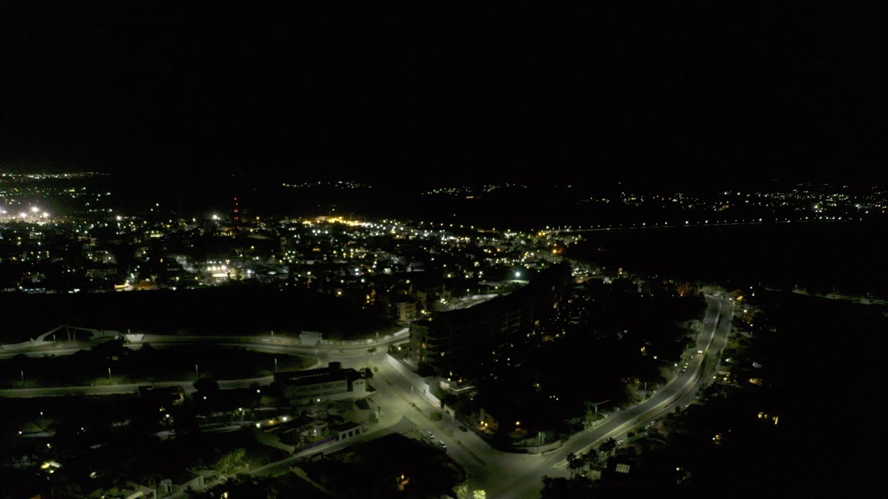 Opening aerial frame over Los Cabos at night, with bright streetlights tracing roads below, distant city lights near a dark horizon, and no visible people.
