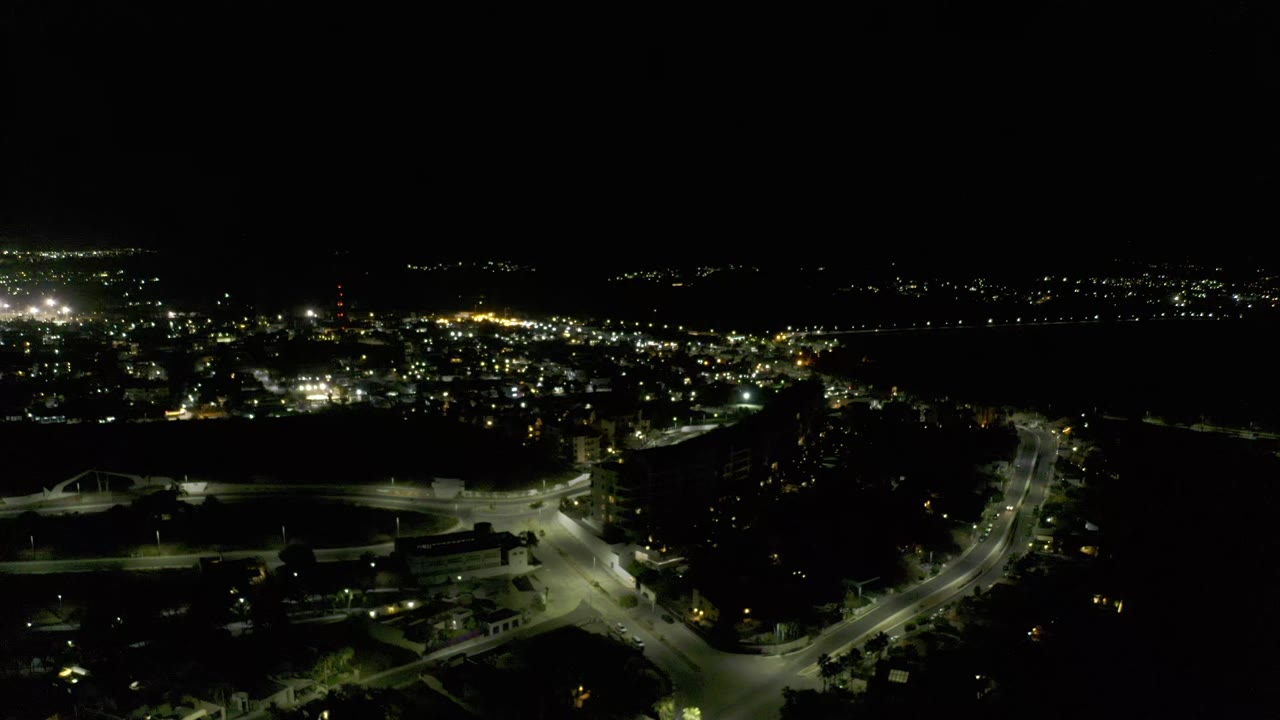 Second aerial frame over Los Cabos, showing a lit intersection and curving road in the lower frame, dense urban lights beyond, and no visible people.