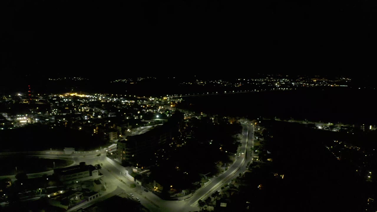 High nighttime view over Los Cabos with a bright road junction in the foreground, clustered dark structures, scattered lights on the horizon, and no visible people.