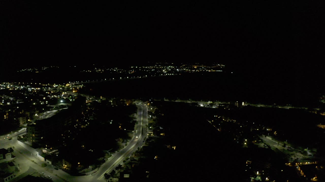 Night aerial view in Los Cabos, Mexico, showing illuminated streets, scattered buildings, distant city lights, and no visible people at the opening moment.