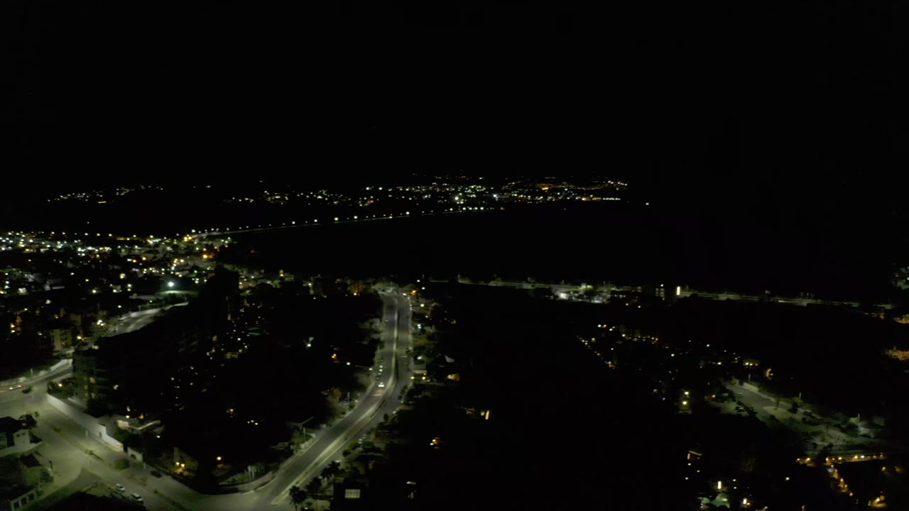 Aerial nighttime view in Los Cabos, Mexico, with a bright curved road in the lower frame, dark building silhouettes, distant lights, and no visible people.