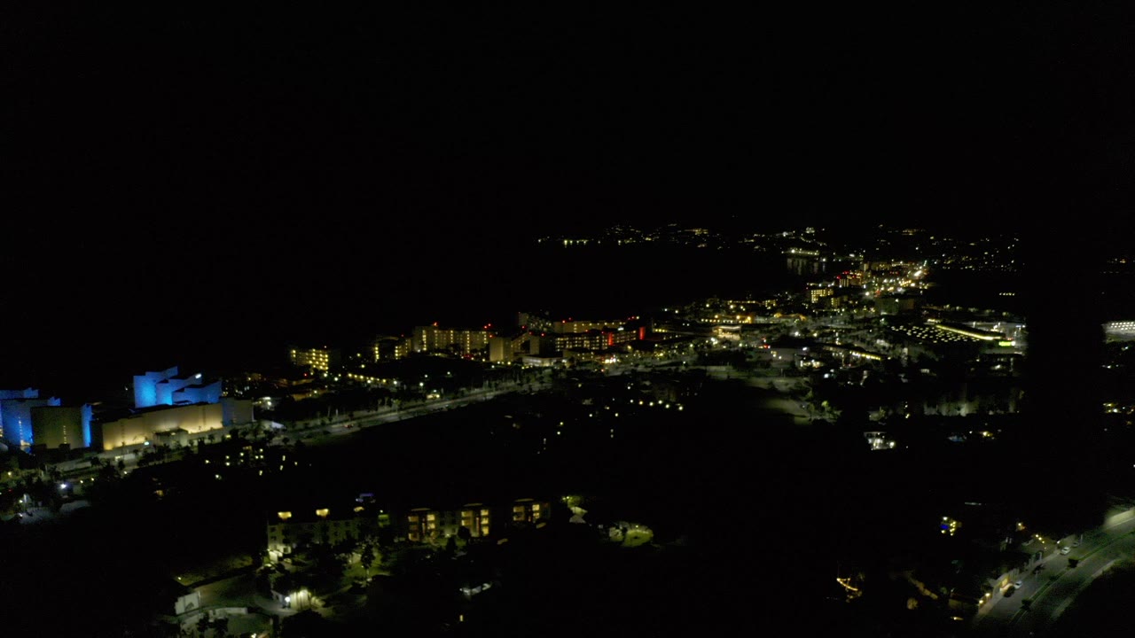 Opening frame of a Los Cabos, Mexico night aerial with a blue-lit building on the left, scattered road lights, distant building clusters, and no visible people.