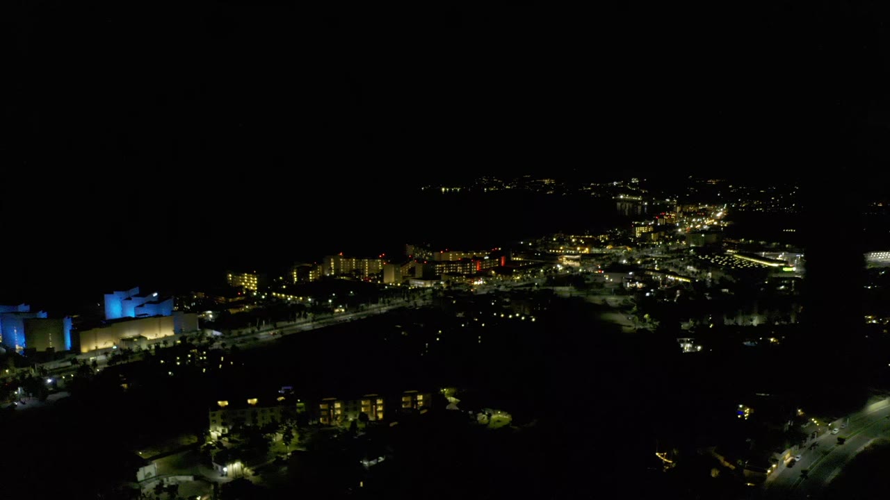 Second frame of the Los Cabos aerial holds on the industrial area, with the blue-lit structure left of frame and dense yellow-white lights across the background, with no visible people.