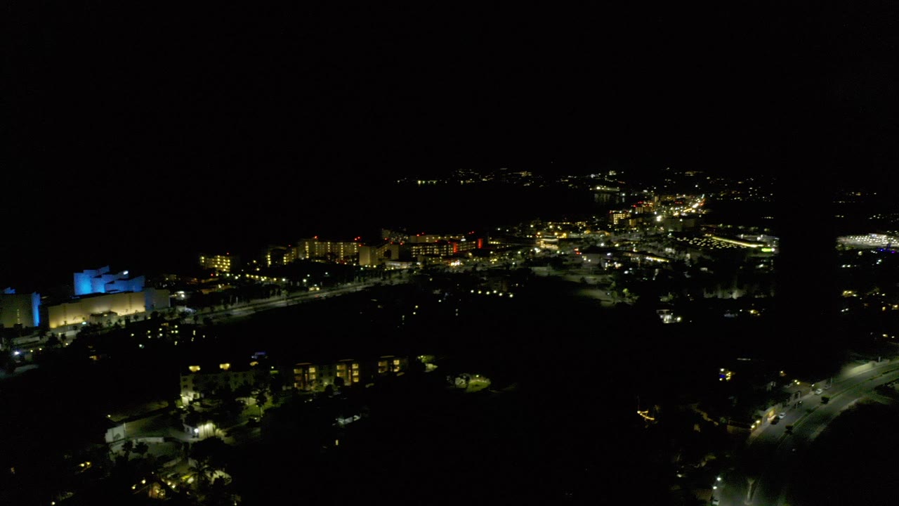 Night aerial view over Los Cabos, Mexico, with scattered street lights, buildings, a blue-lit structure at left, and no visible people.