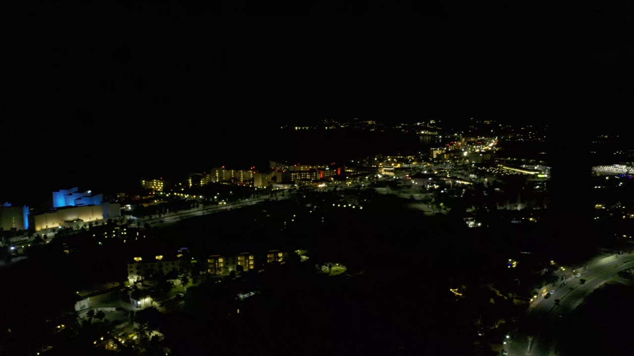 Los Cabos nighttime aerial frame with bright road lines crossing the lower right, clustered buildings near the center, and no visible people.