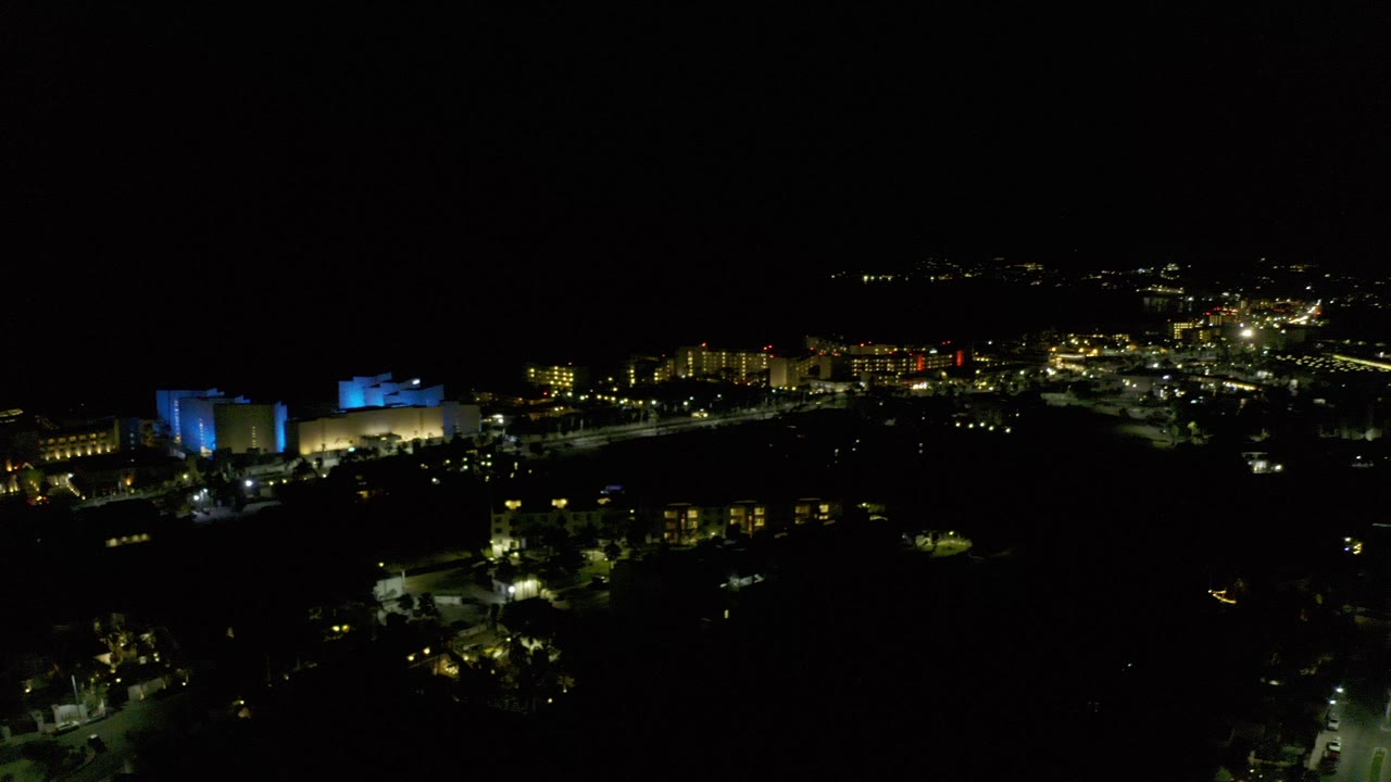 Opening frame of Los Cabos at night from a high aerial view, with a blue-lit building cluster at left, scattered yellow city lights, dark foreground, and no visible people.