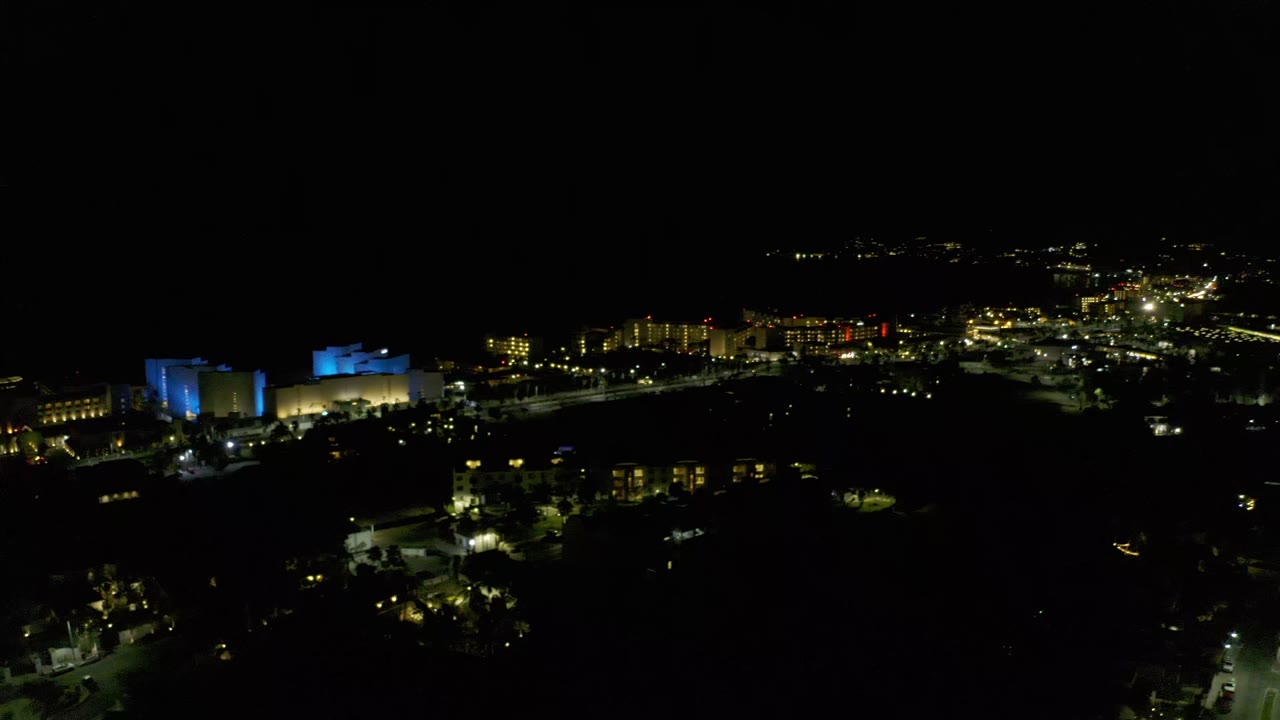 Los Cabos night skyline seen from above, with bright road lights spreading across the right side, blue-lit buildings left of center, and no visible people.