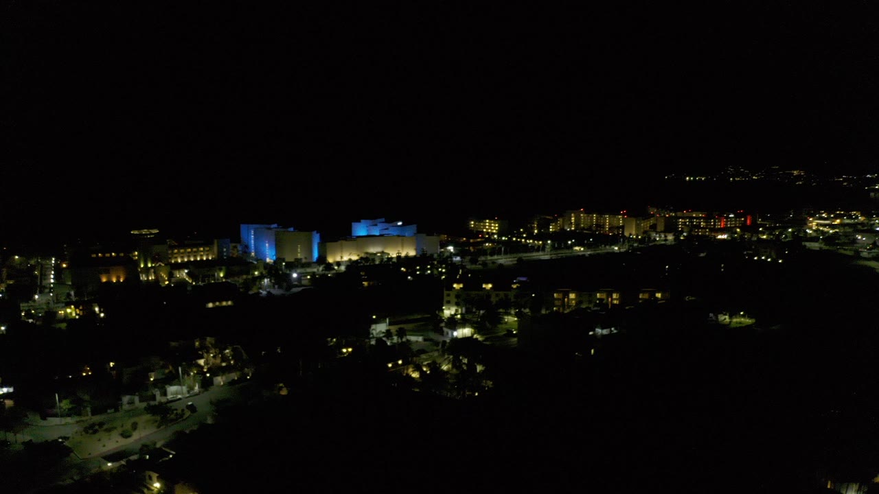 Wide dark night view over Los Cabos, Mexico with scattered yellow and blue city lights, dark foreground, and no visible people.