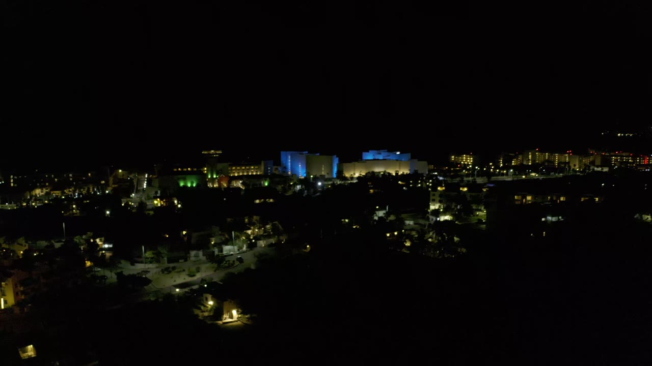 Los Cabos night skyline seen from an elevated distance, with blue lit structures near center, yellow window lights to the right, scattered streetlights below, and no visible people.