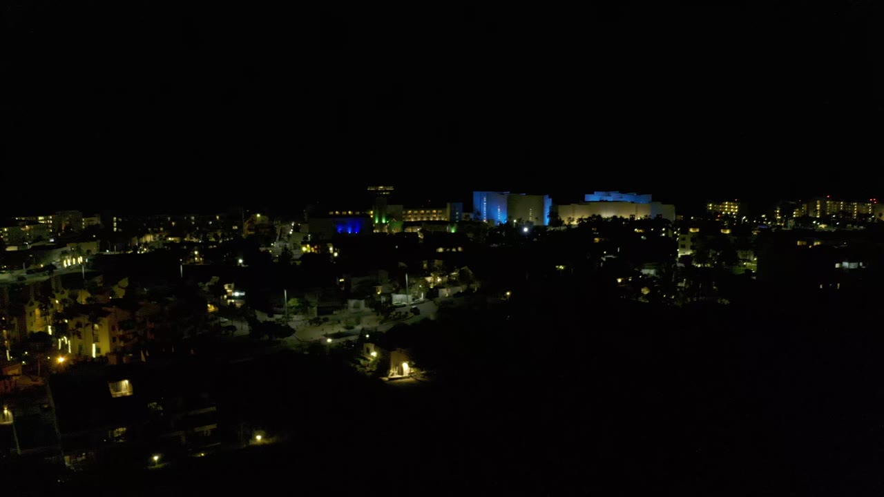 Second frame shows the Los Cabos night skyline slightly farther across the same view, with illuminated blocks, a blue-lit structure near the horizon, and no visible people.